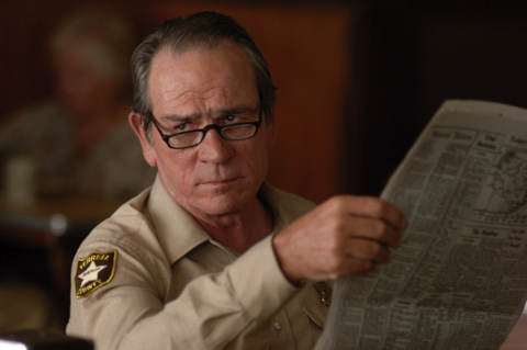 Tommy Lee Jones holding a newspaper, looking just past the camera at a character in a way that implies they said something really stupid.