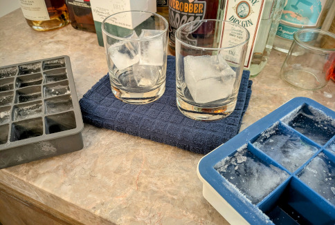 two rocks glasses with cube-shaped ice, each sitting next to its corresponding ice mold. The glass on the left has three smaller half-clear cubes, and the mold's yield is 15. The glass on the right contains one large cube that's partially clear, and the mold's yield is 6.