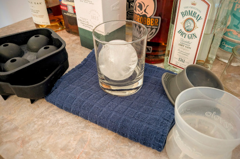 a rocks glass with a large, half-clear ice sphere. On its right is the single-yield plastic mold matching the sphere, with a bottom 'cup' and a removable half-sphere rubber top. To the left of the glass is another empty mold, with a tray and a lid, that can make four spheres