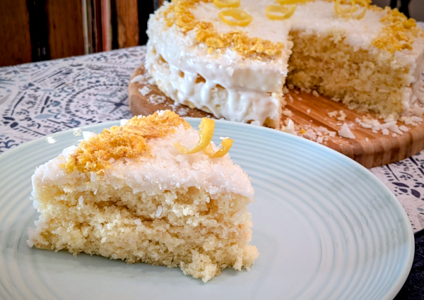 a triangular slice of yellow cake with white, coconut-dusted icing, topped with a strip of crushed plantain chips and a spiral twist of lemon peel; the rest of the cake sits in the background