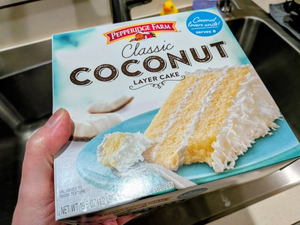 Skep holding the box of cake in one hand; the box measures maybe 8 inches to a side. The box is labeled “Pepperidge Farm Classic Coconut Layer Cake” with a blurb in the corner that reads “Coconut lovers unite! serves 8”. The image on the box shows a slice of cake, with three layers of yellow sponge and white icing topped with shavings of coconut.