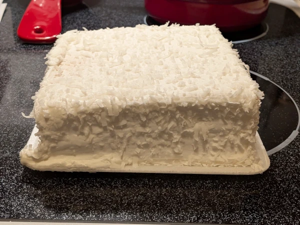 The cake is now removed from the box and sits on the stove. It is a square cake, covered in icing and coconut shavings as the box depicts, though in a more underwhelming way. The left side of the cake seems to be sagging.