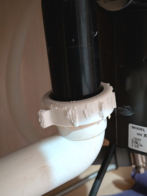 A photo of a white connector between two pipes, taken in the cabinet below the sink. The plastic connector is damaged and mangled.