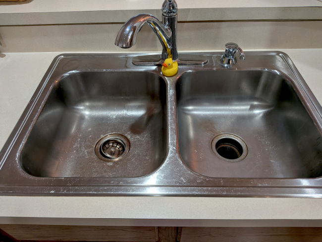 A photo of a stainless steel two-bowl sink with a rubber duck perched on the center divider.