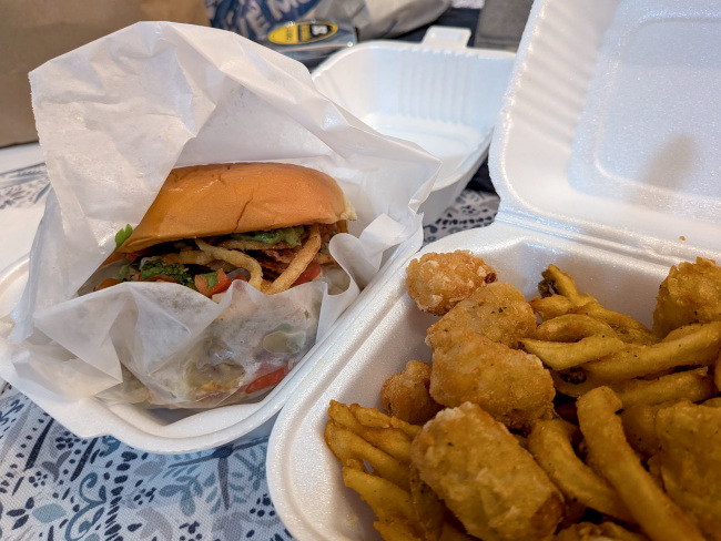 A photo of a take-out burger with toppings and a mix of tater tots and french fries.