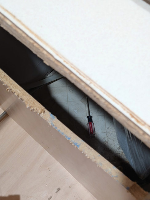 A photo from above the counter looking downward through the sawed-out piece of cabinet wall, showing a dropped screwdriver laying in the empty space between the sink cabinet and the dishwasher.