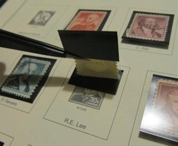 A photo of a stamp collection page; various stamps are already mounted. A stamp in the center is currently in the process of being mounted; tweezers are holding up the bottom of the stamp, showing how it is resting in the lower fold while the upper fold is affixed to the page.