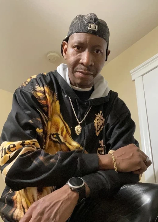 A photo of a man kneeling over the camera, wearing the black lion-print hoodie, black pants, backwards ball cap, digital watch, gold chain bracelet, and gold chain necklace with a lion’s head pendant.
