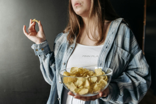 a photograph of a woman in loose-fitting clothes holding a bowl of potato chips in one hand and a single chip in the other