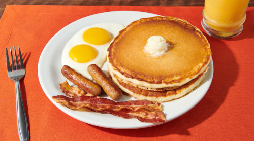 a photograph of a plate containing pancakes with a pat of butter, eggs sunny-side-up, two sausage links, and two slices of bacon, with a fork and glass of orange juice nearby