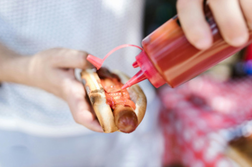 a photograph of a person appling ketchup of all condiments to a sad-looking hot dog