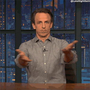 a gif of Seth Myers from Late Night with Seth Myers sitting at his desk facing the camera and gesturing around himself